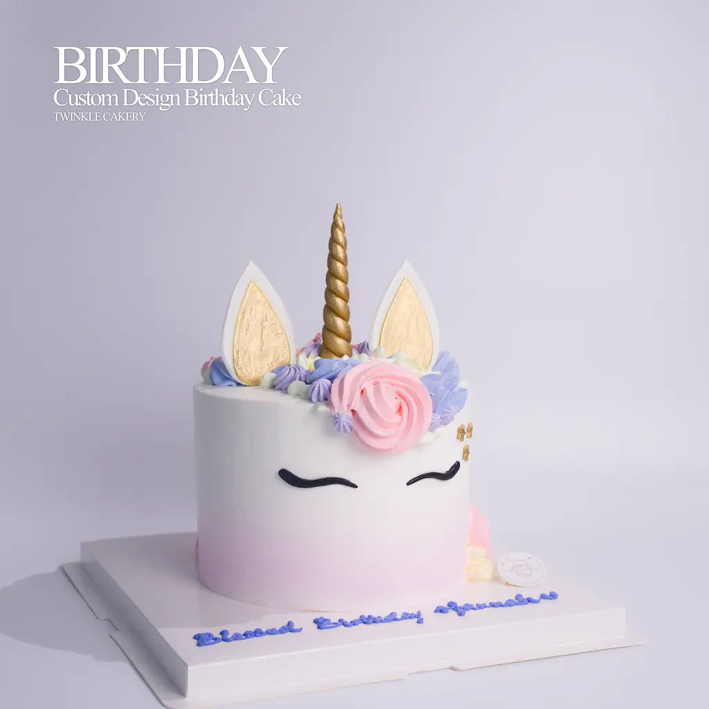 A custom unicorn birthday cake from Twinkle Cakey, featuring a white to light pink gradient base, gold horn, white and gold ears, pink and blue frosting flowers, and closed-eye decoration, presented on a white cake board with 'Blessed Birthday Aummi!' written in blue icing. By Twinkle Cakery. Delivery across Selangor & KL

